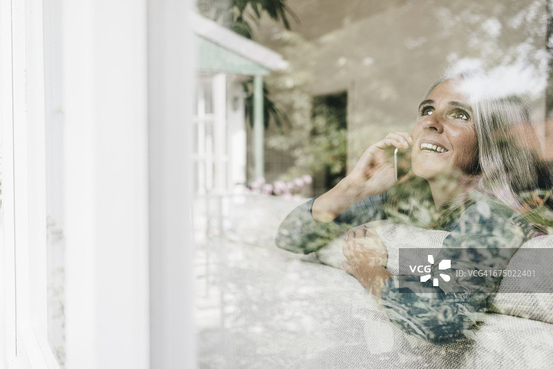 微笑的女人坐在沙发玻璃窗后面看电话图片素材