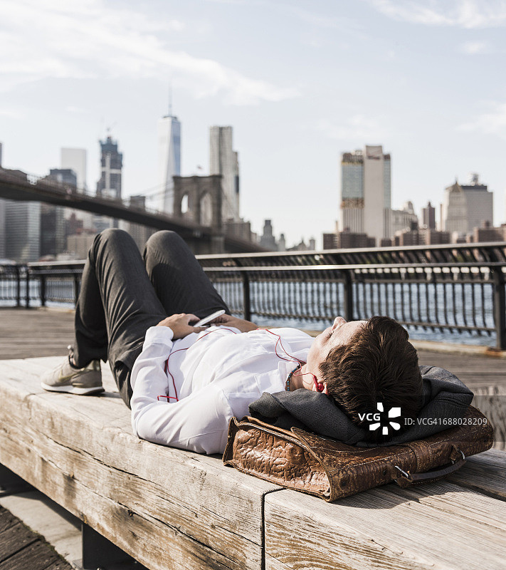 美国，布鲁克林，女商人躺在长凳上用耳机听音乐图片素材