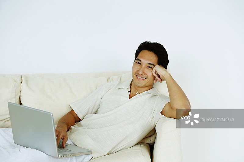一个男人躺在沙发上，用笔记本电脑，手放在头上图片素材
