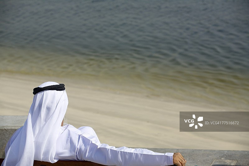 阿拉伯人坐在公园长椅上，看着水。阿拉伯联合酋长国迪拜图片素材