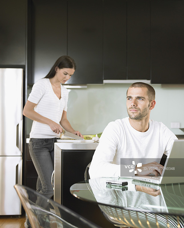 女友切菜时，男人拿着笔记本电脑坐在桌边图片素材
