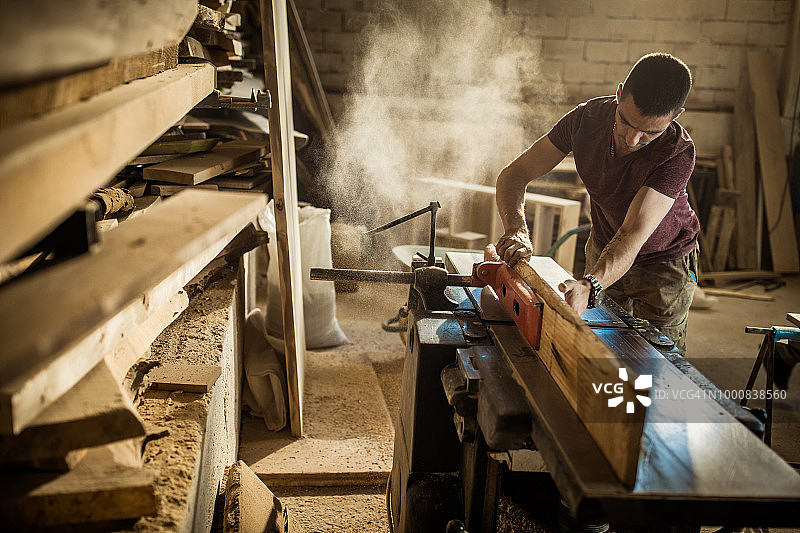 一个年轻的男木匠在车间里的一块木头上工作。图片素材