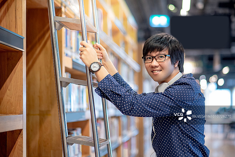 年轻的亚洲男子学生在公共图书馆的书架上使用梯子选择书籍，男性研究员戴着耳机和眼镜在大学学院教育和学习概念做研究图片素材