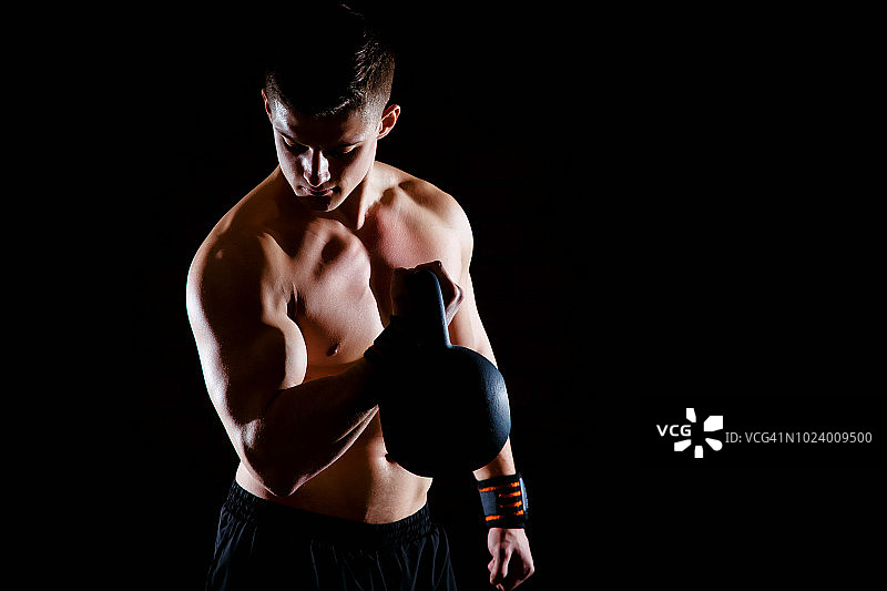 在黑暗的背景下，健康的肌肉发达的年轻人拿着哑铃做重量锻炼。图片素材
