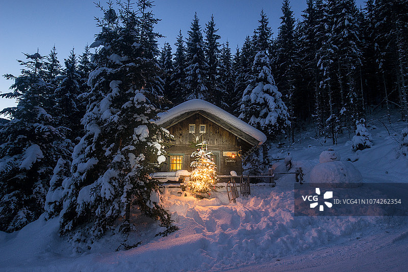 在奥地利的Altenmarkt-Zauchensee，雪橇，雪人和圣诞树在夜晚的雪中照亮了木屋图片素材