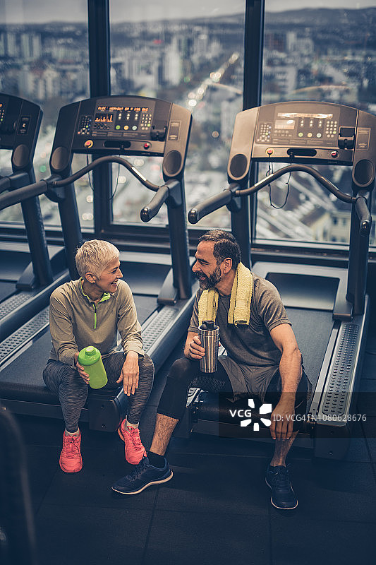 一对快乐的老年夫妇在健身俱乐部的跑步机上休息，互相聊天。图片素材