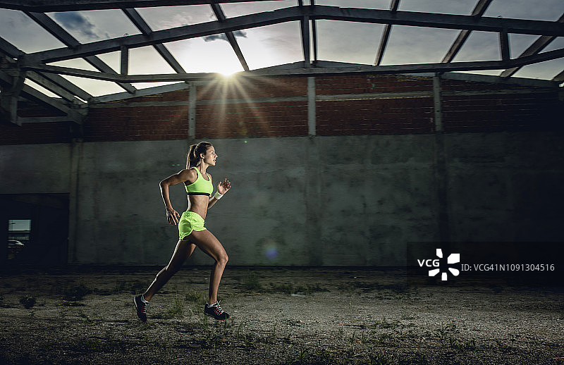 一个女运动员在旧仓库跑步的侧面图。图片素材