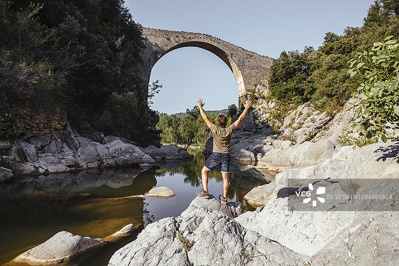 在阳光明媚的日子里，站在岩石上的人举起手臂看着拱桥的背影图片素材