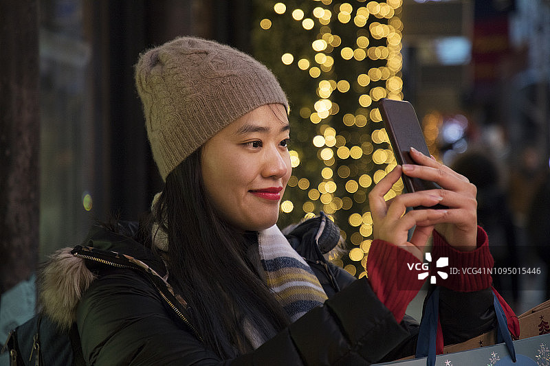 一名亚洲妇女在城市街道上用手机拍摄圣诞装饰品。图片素材