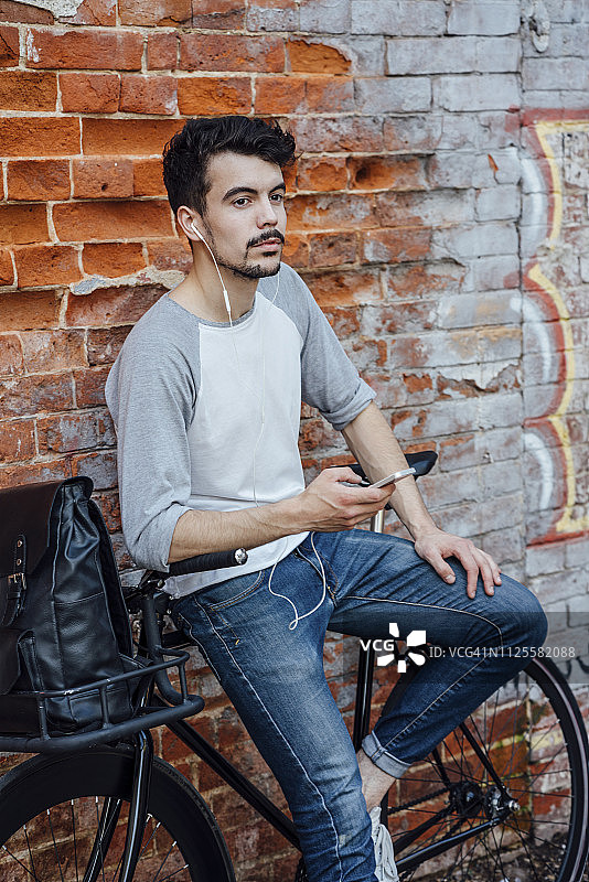 一个年轻人骑着自行车，站在砖墙前，手里拿着手机和耳机图片素材