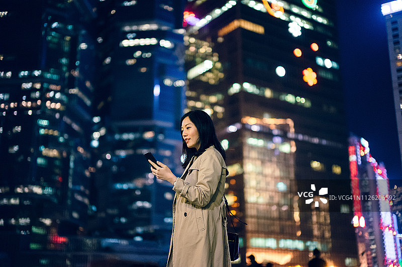 自信的年轻女商人用智能手机对抗香港金融区的现代摩天大楼图片素材