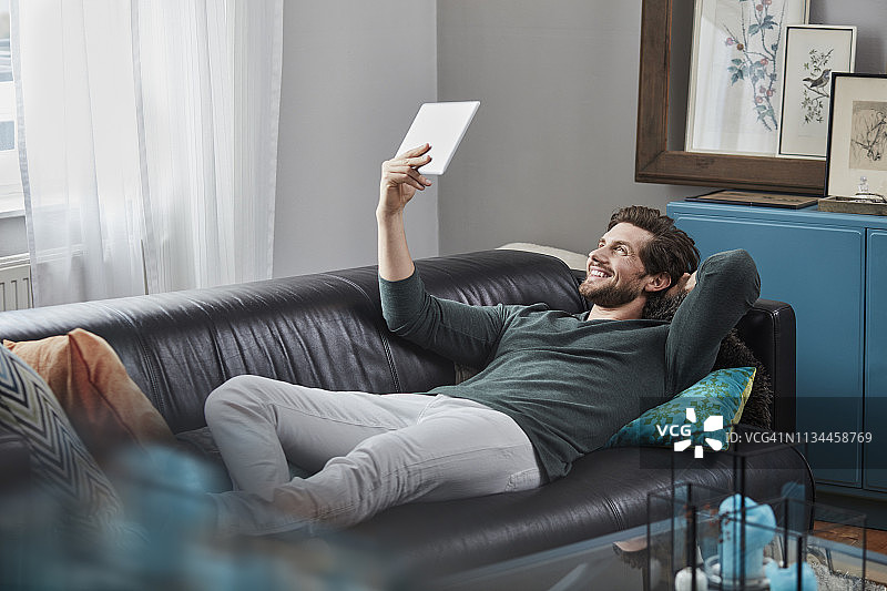 一个面带微笑的男人躺在家里的沙发上，拿着平板电脑图片素材
