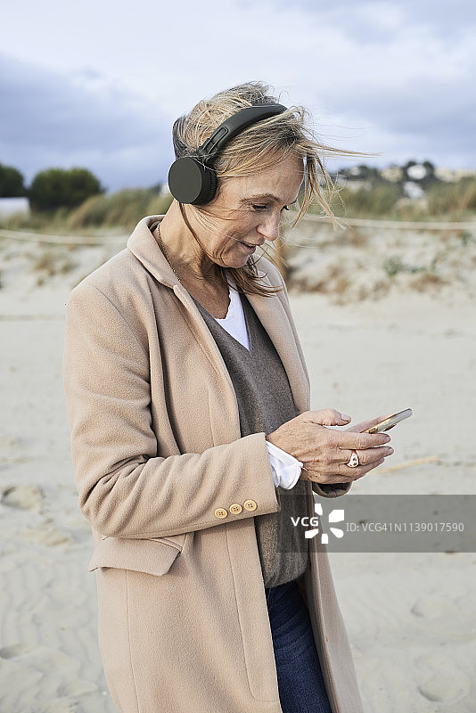 西班牙的梅诺卡，一名老年女性在冬天的海滩上使用智能手机和无线耳机图片素材