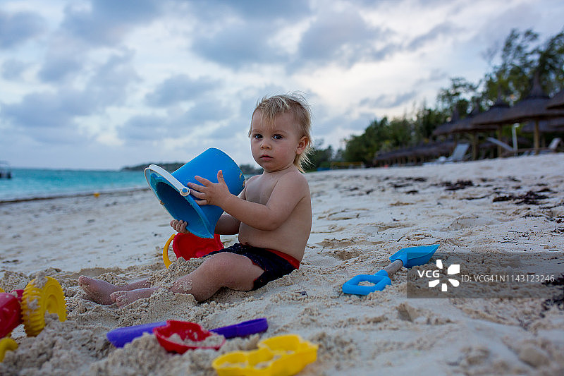 可爱的蹒跚学步的小男孩在热带海滩上玩海滩玩具图片素材