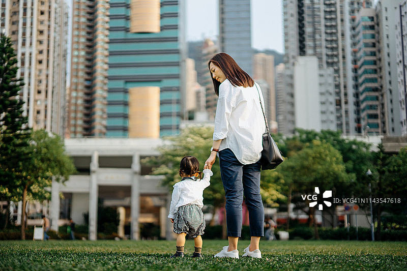 年轻的母亲和女儿手牵着手享受美好的一天，在现代城市的天际线的城市公园散步图片素材