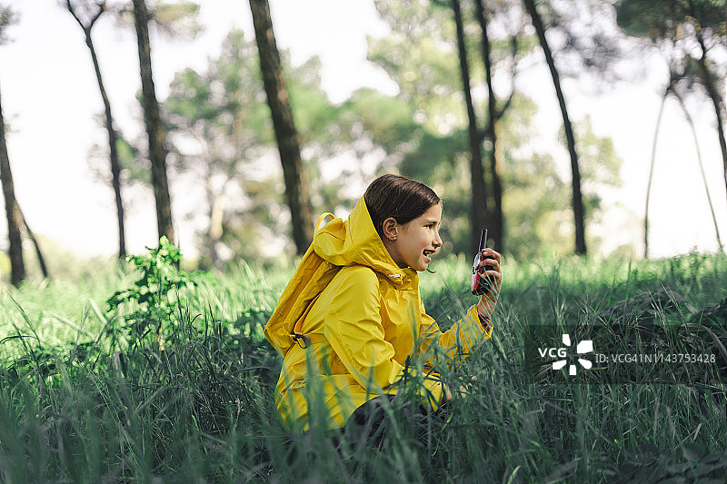 穿着黄色雨衣，背着黄色背包，拿着对讲机的女孩蹲在草地上图片素材