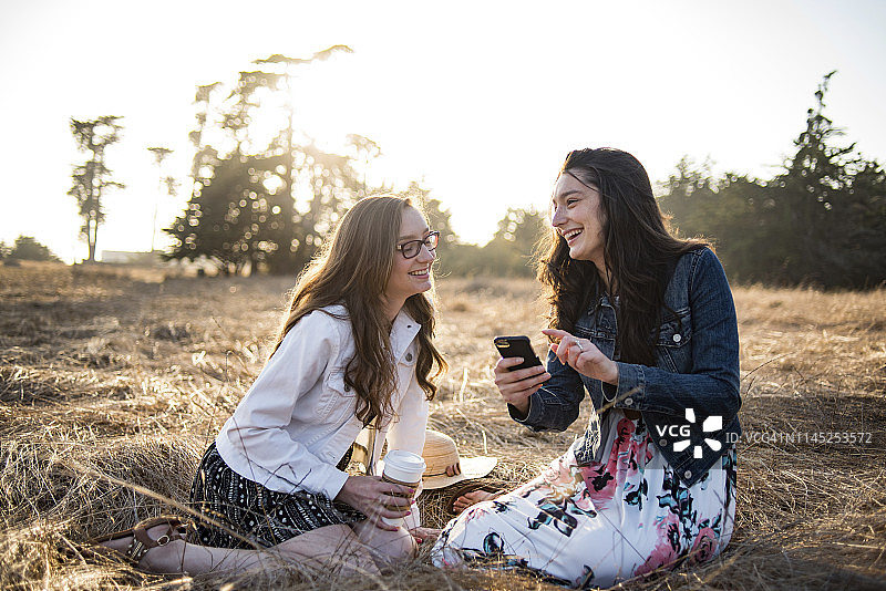 快乐的姐妹们坐在草地上玩智能手机，日落时天空晴朗图片素材