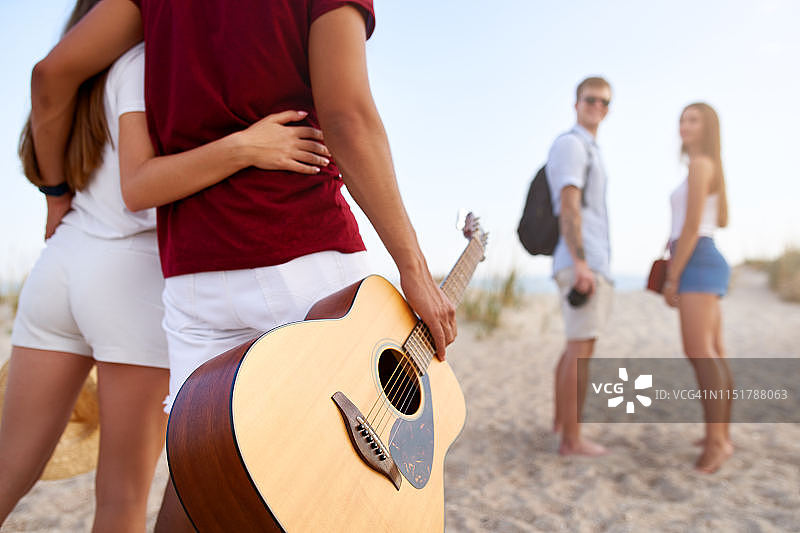 日落时分一群朋友去沙滩。年轻的男男女女走到海边，在海边放松，弹吉他。青少年在假期。男朋友和女朋友拥抱。孤立的观点图片素材