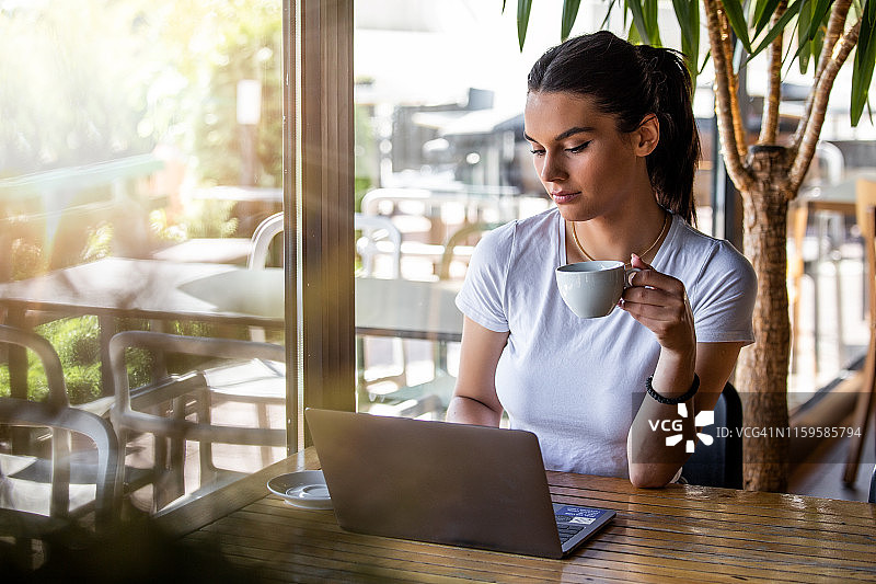 一个年轻漂亮的女孩坐在靠窗的咖啡馆里，喝着卡布奇诺咖啡。在咖啡店用笔记本电脑工作的漂亮女人。有魅力的女人坐在咖啡馆里拿着笔记本电脑图片素材