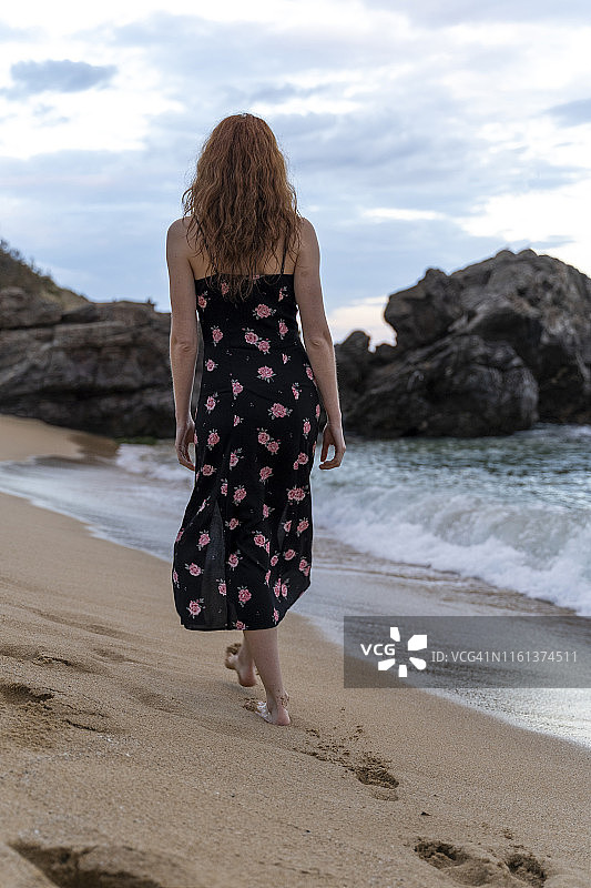 后视图的年轻女子穿着夏天的连衣裙与花设计walkung在海滩上图片素材