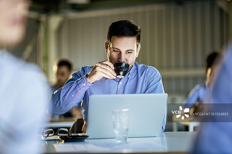 年轻的商人一边喝咖啡一边在满是人的办公室里用笔记本电脑工作。图片素材