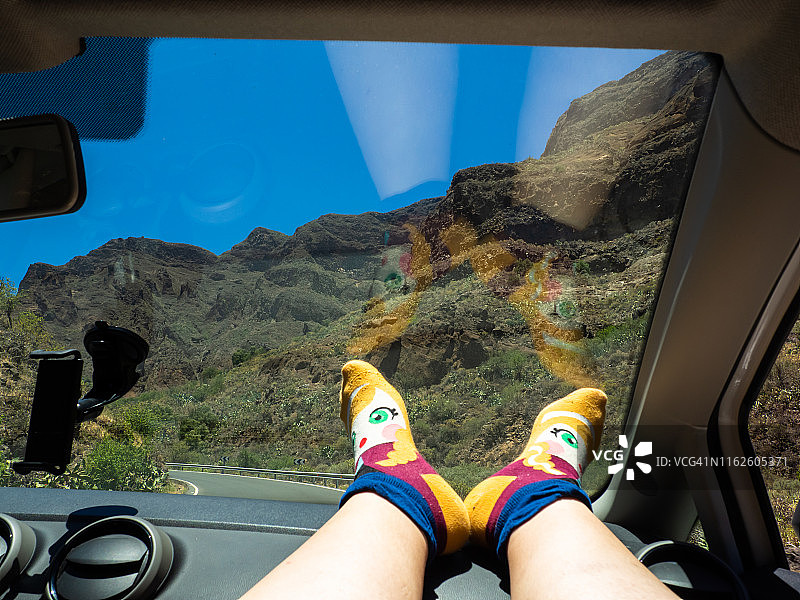 一个穿着滑稽袜子、脸上涂着油彩的女人的脚停在一辆行驶在路上的汽车的仪表盘上图片素材