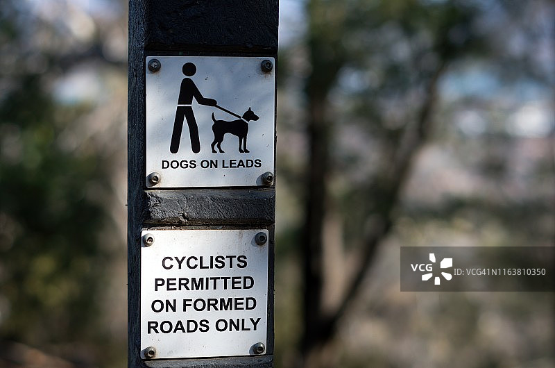 在澳大利亚首都地区堪培拉的安斯利山自然保护区，一条小路上的指示牌上写着“狗狗在引路”和“自行车只能在路面上骑行”图片素材