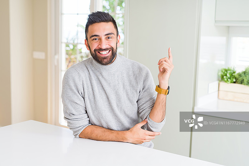 英俊的西班牙裔男子穿着休闲毛衣在家带着大大的笑容，用手指着手指一边看着镜头。图片素材