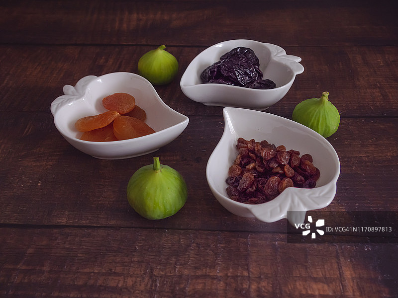 秋天的水果:一簇簇的杏干、梅干和葡萄干放在心形的瓷碗里，新鲜采摘的绿色无花果放在旧木桌上图片素材