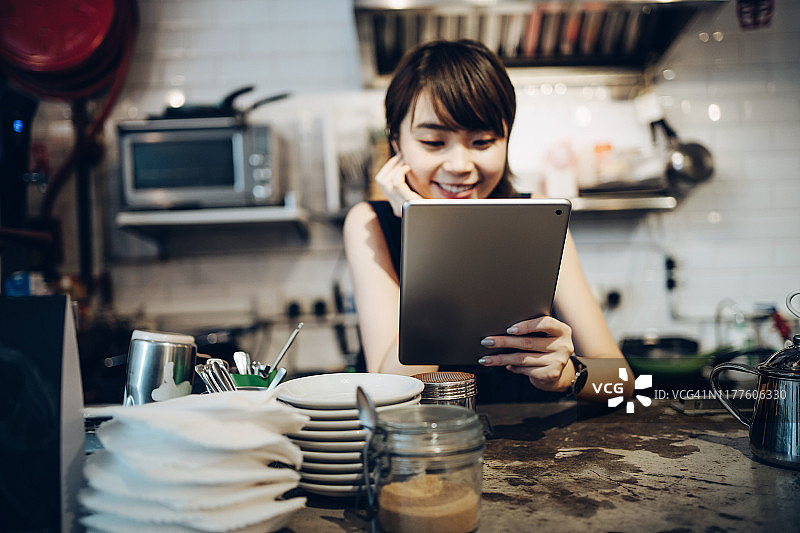 年轻的亚洲女咖啡师一边在咖啡店吧台后面工作，一边使用着数字平板电脑图片素材