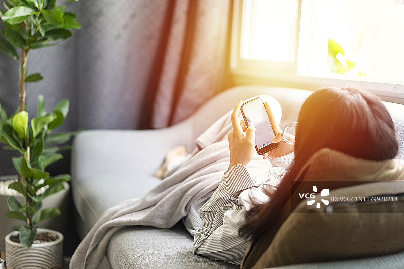 美丽的亚洲女孩女性在舒适的沙发上玩智能手机与空气净化器图片素材