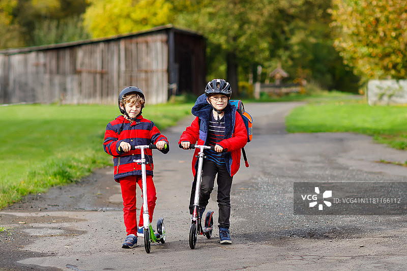 两个小男孩骑着滑板车上下学。7岁的小学生开车穿过雨坑。有趣的兄弟姐妹和最好的朋友一起玩。儿童学校。图片素材