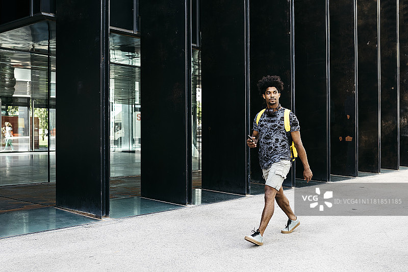 西班牙巴塞罗那，一名背着黄色背包、带着智能手机和耳机的男子走在大街上图片素材