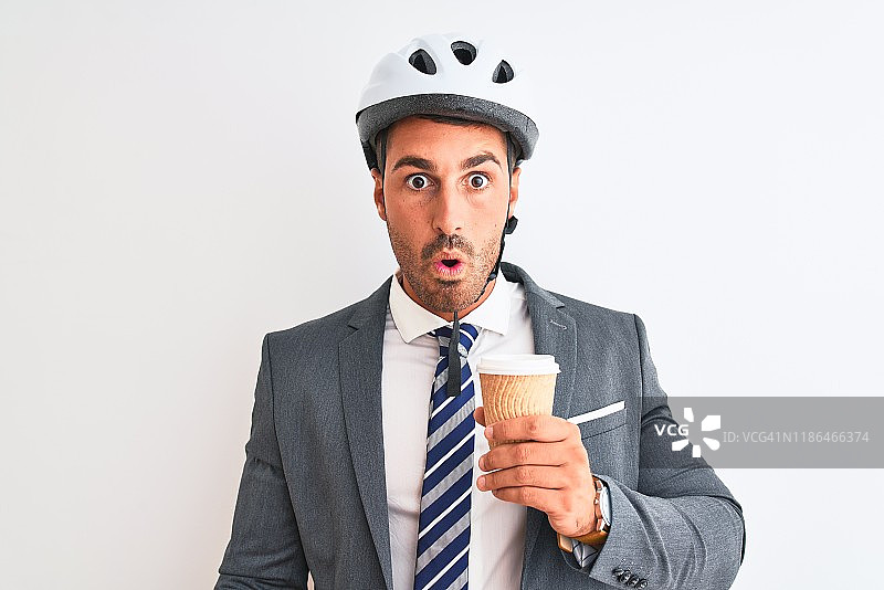 年轻英俊的商人戴着自行车头盔，带着咖啡走过孤立的背景，惊恐中带着惊讶的表情，害怕中带着兴奋，害怕中带着恐惧的表情图片素材