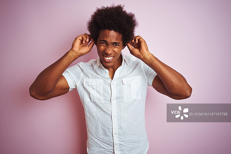 年轻的美国男子与非洲式头发穿白衬衫站在孤立的粉红色背景覆盖耳朵与手指与恼怒的表情噪音的音乐。聋的概念。图片素材