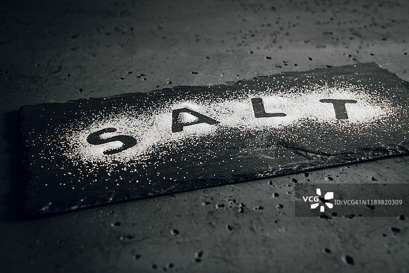 把白色的厨房盐洒在石头桌上。盐放在桌子上，上面刻着“盐”的字样。白盐对身体有害的观念，高血压的问题。图片素材