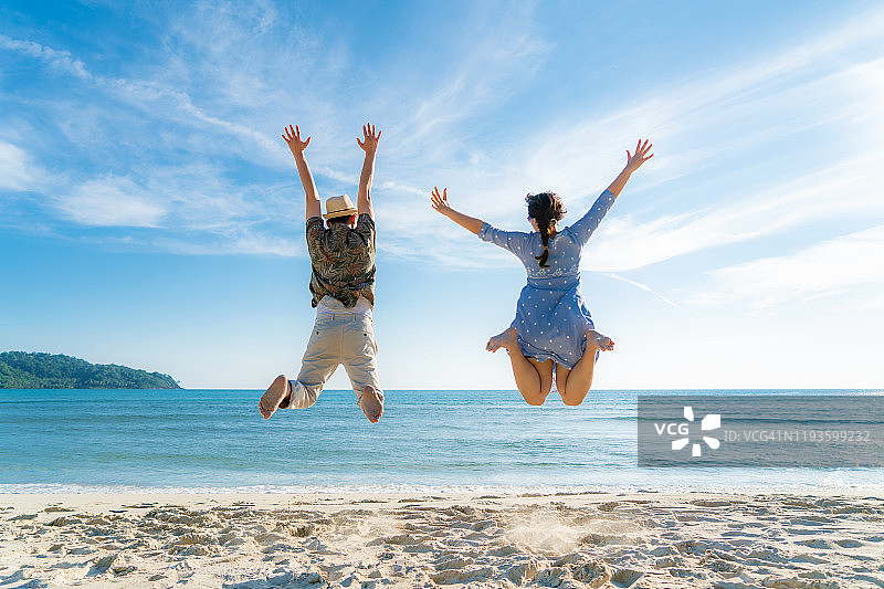 快乐的亚洲夫妇游客跳在海滩度假。在完美的白色沙滩和阳光下的热带海滩上，年轻夫妇为夏日假期欢呼，展示成功、幸福和快乐的旅游理念。图片素材