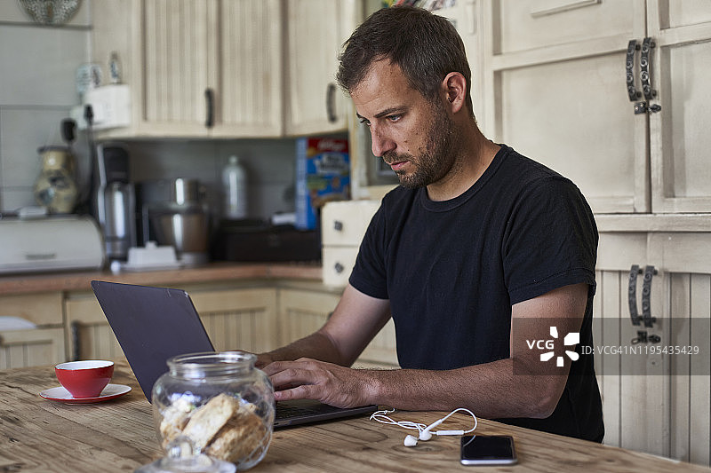 男人在家工作，坐在餐桌前，使用笔记本电脑和智能手机图片素材