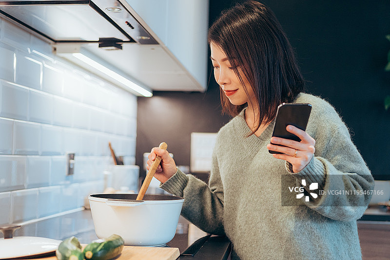 年轻快乐的女人拿着智能手机在家厨房做饭图片素材