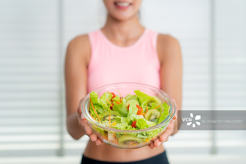一个穿着粉红色运动服的快乐苗条女人的肖像，上面显示着家里厨房里的一碗新鲜蔬菜沙拉。健康的饮食和生活方式为苗条和良好的健康理念图片素材