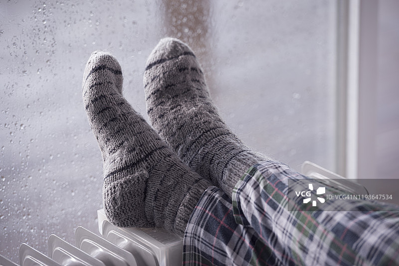 女人的脚上穿着灰色的羊毛袜，在雨冬季节用油加热器暖脚。图片素材