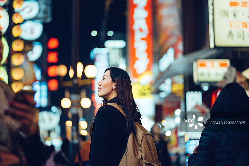 微笑的年轻亚洲女性游客探索和漫步在繁忙和五颜六色的霓虹灯招牌的市中心街道在日本大阪的夜晚图片素材