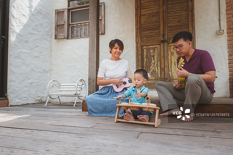 快乐时刻年轻的亚洲家庭观念，母亲和父亲享受弹奏尤克里里，小儿子打鼓时坐在房子前，家人放松和一起在家里度过时间图片素材