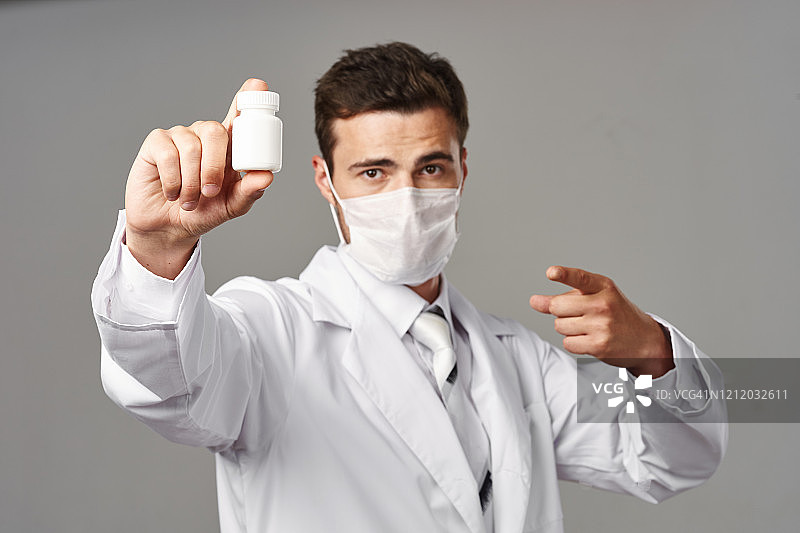 一名身穿白大褂、面带医用口罩的经验丰富的医生展示了一罐针对病毒冠状病毒流行的药物，背景为灰色。图片素材