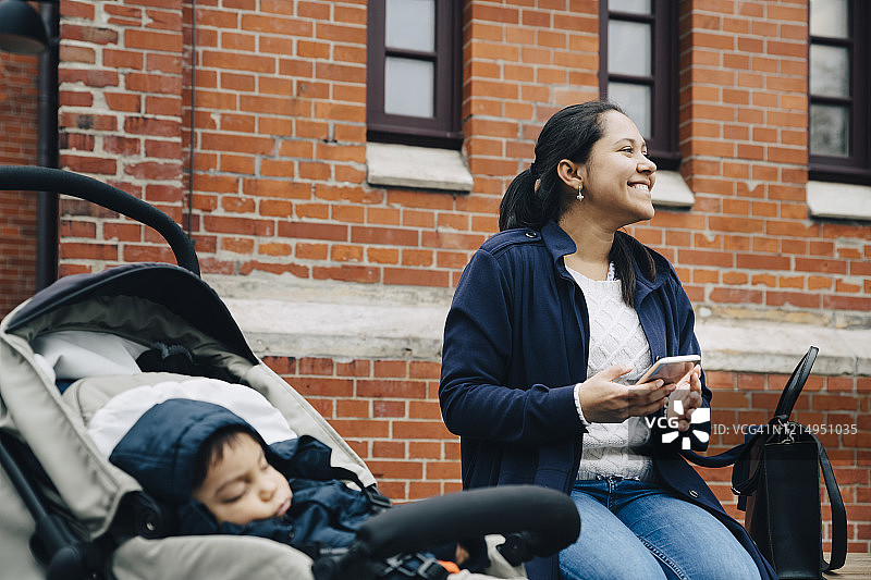微笑的母亲使用智能手机，而儿子坐在婴儿车对着城市的建筑图片素材