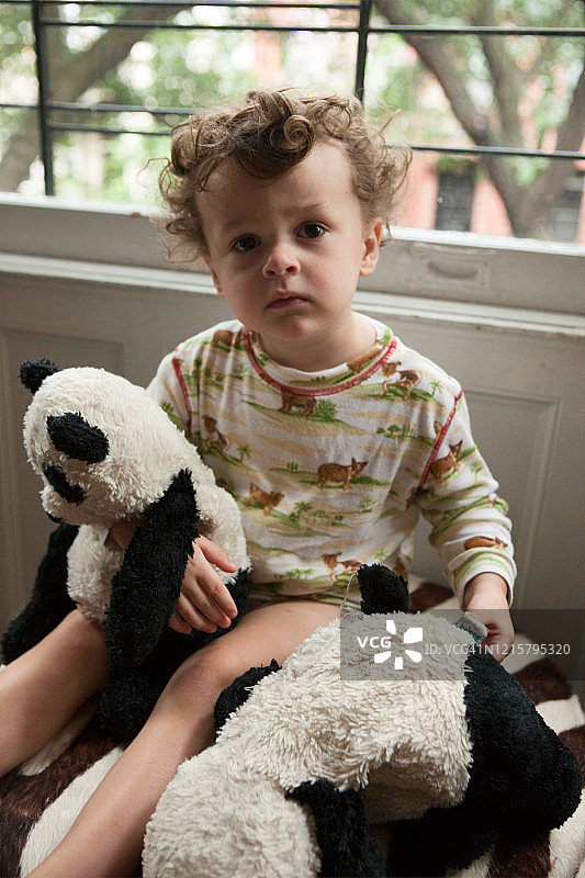 穿着睡衣蹒跚学步的小男孩抱着两只毛绒熊猫，坐在窗前。图片素材