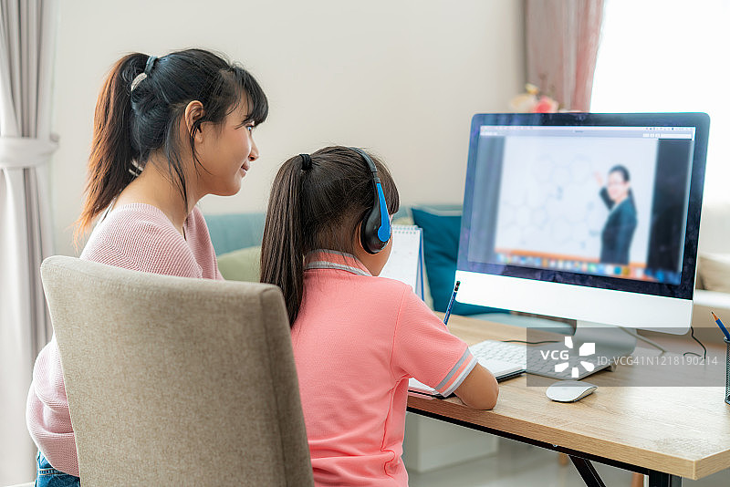亚洲女学生与母亲视频会议电子学习与教师在电脑上在家里的客厅。在家教育和远程学习，在线，教育和互联网。图片素材