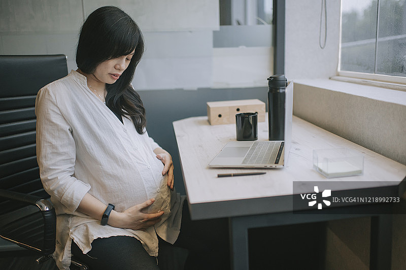 一位亚裔怀孕白领在她的办公室隔间里休息，和她肚子里的双胞胎宝宝聊天图片素材