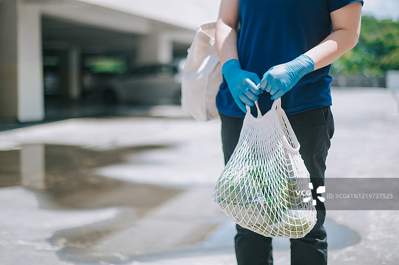 一位亚裔华人中年妇女带着从超市买的食品杂货走在停车场里，戴着医用橡胶手套图片素材