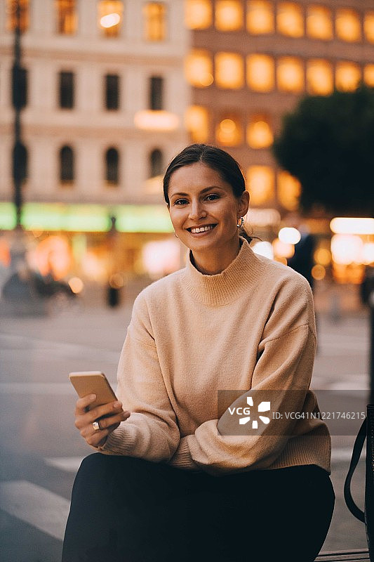 微笑的年轻女子坐在城市里使用手机的肖像图片素材